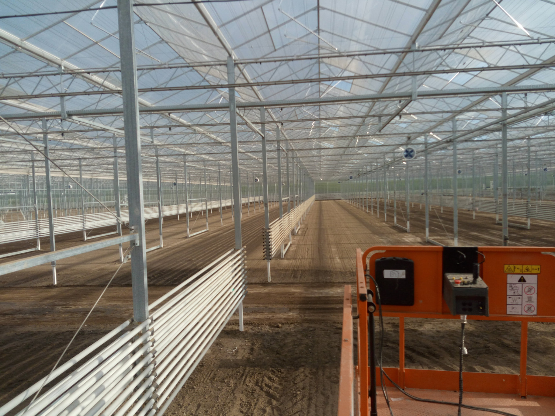 Nieuw gebouwde kas met stalen draagconstructie, rijen witte verwarmingsbuizen en een hoogwerker op de losse grond